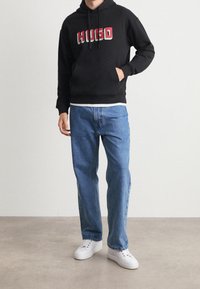 Felpa nera con logo "HUGO" in rosso e verde, abbinata a jeans dritti azzurri e sneakers bianche su uno sfondo neutro.