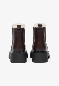 Bottines en cuir marron avec un col en fausse fourrure, semelle noire épaisse et détail en patte à l'arrière. Texture lisse avec coutures visibles.