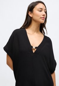 Črna bluza z V-izrezom in kratkimi rokavi. Opremljena z okrasno zlato zaponko na vratu in teksturiranim zaključkom tkanine.