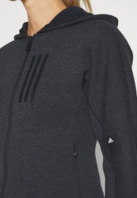 Haut à capuche noir avec fermeture éclair, fabriqué en tissu doux. Caractéristiques de trois bandes audacieuses sur la poitrine et d'un écusson logo sur la manche.