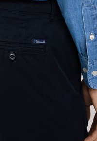 Navy blauwe broek met een gladde textuur, voorzien van een achterzak en een klein wit label met "Faconnable" erop gestikt. Knopsluiting zichtbaar.