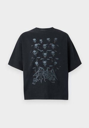 Άνδρας που φοράει μαύρη φαρδιά μπλούζα με σχέδιο κρανίου, γκρίζο παντελόνι με φαρδιά πόδια, μαύρα χοντρά παπούτσια, σκούρα γυαλιά ηλίου και ασημένιο κολιέ με αλυσίδα.