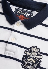 Polo blanc à rayures bleu marine, avec deux boutons et un col bleu marine. Présente un blason brodé avec l'inscription « Superdry » et la date "1954".