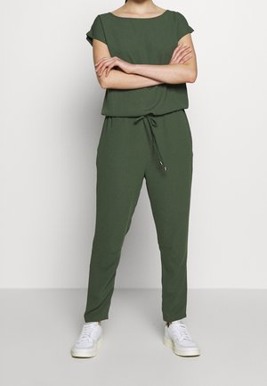 Kvinde står med armene over kors iført olivengrøn buksedragt og hvide sneakers mod en ensfarvet hvid baggrund.