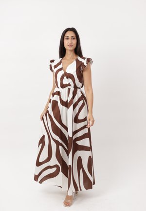 Mujer de pie que lleva un vestido maxi blanco sin mangas con un patrón abstracto de remolinos marrones y detalles de volantes en los hombros.