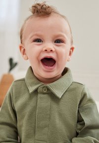 Zaļš teksturēts krekls ar apkakli un pogu aizdari, garām piedurknēm ar kontrastējošu paneli.