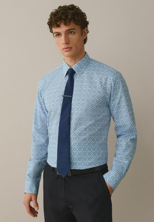 Junger Mann trägt ein hellblaues Hemd mit floralem Muster, eine marineblaue Krawatte und schwarze Hose, die Hand in der Tasche, neutraler Hintergrund.