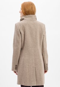 Donna con capelli rossi che indossa un cappotto di lana beige con colletto alto, vista da dietro su uno sfondo bianco uniforme.