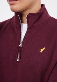 Burgundské zipové mikina s vysokým límcem, s kovovým stříbrným zipem a malým žlutým vyšitým ptáčkem na hrudi.