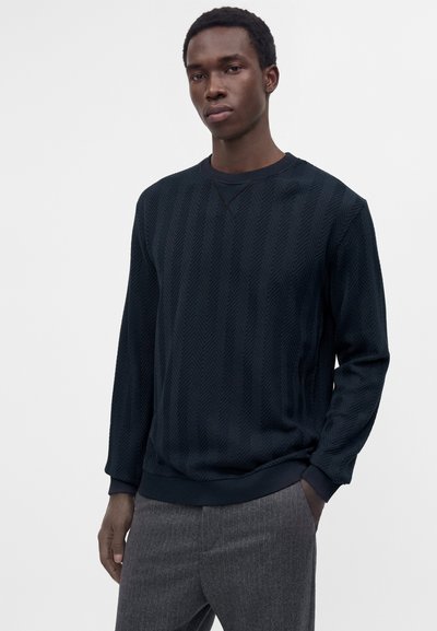 Jeune homme à la peau foncée portant un pull noir texturé à manches longues et un pantalon gris, debout avec une main dans la poche sur un fond uni.