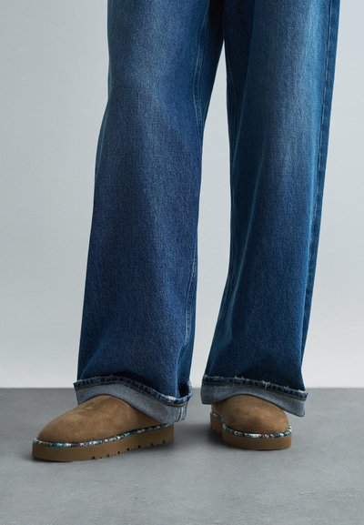 Hnědé semišové nazouvací boty s texturovaným svrškem, které mají multikulturní tkaný prvek kolem podrážky, kombinované s širokými modrými džíny.