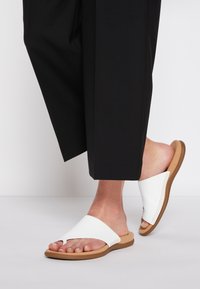 White slide sandals with a wide strap and open toe, featuring a tan sole and decorative stitching along the edge. Black pants partially visible.