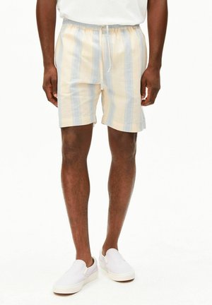 Mann trägt beige und hellblaue Shorts mit vertikalen Streifen, Kordelzugbund und weißen Slip-on-Schuhen, steht vor weißem Hintergrund.