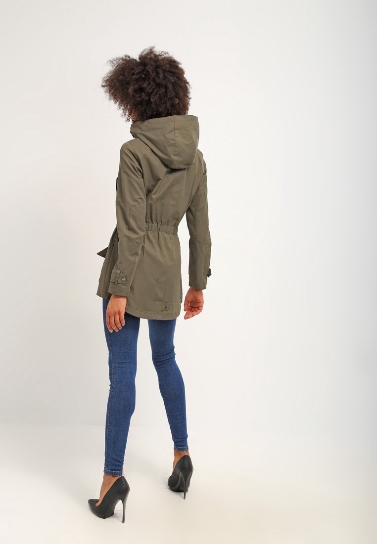 Olivově zelený, lehký parka s kapucí, stahovaným pasem a bočními kapsami, doplněný modrými skinny džíny a černými vysokými podpatky.