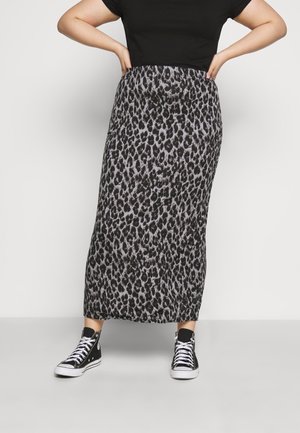 Persoon die een zwart shirt, een enkellange rok met luipaardprint en zwarte hoge sneakers draagt, met de handen op de heupen staand tegen een effen achtergrond.