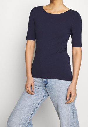 Personne portant un haut bleu marine ajusté à manches courtes et un jean taille haute bleu clair, debout contre un fond uni.