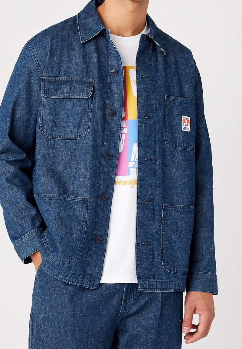 Farkkutakki, tummansininen, jossa on neljä taskua, nappi eteen ja kaulus. Päällä valkoinen graafinen t-paita, jossa on värikäs kuvio.