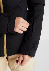 Svart isolerad jacka med guldzipper, som har en stilren design och en ficka med dragkedja i ärmslutet för säker förvaring.