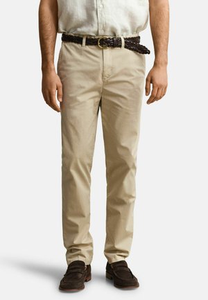 Homme portant un pantalon chino beige, une ceinture tressée marron foncé et des mocassins marron foncé, debout avec les mains détendues le long du corps.