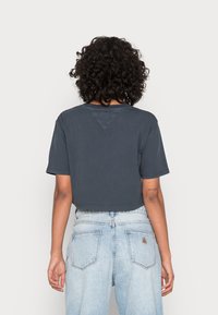 Zkrácená tmavě modrá tričko s krátkými rukávy, s kulatým výstřihem a trojúhelníkovým panelem na zadní straně. Kombinováno se světle modrými džíny.