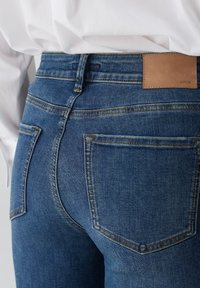 Jean en denim bleu foncé avec une étiquette en cuir tan portant la mention "OPUS". Ses caractéristiques comprennent des poches arrière courbées et des surpiqûres contrastées visibles.
