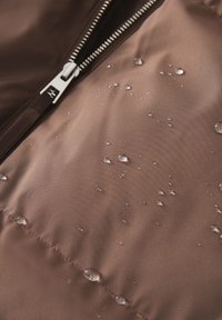 Tessuto di giacca impermeabile marrone con gocce d'acqua. La vista ravvicinata mostra la texture liscia e una cerniera argentata con design a denti.