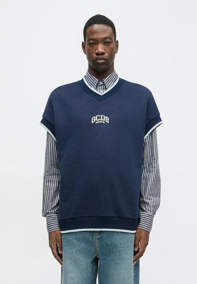 Sweatshirt oversized azul-marinho com decote em V, apresentando detalhes azul claro e o logo "GCDS". Usado por cima de uma camisa com gola azul e branca listrada.