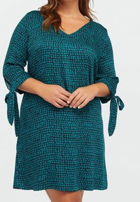 En teal och svart tunikaklänning med V-ringning, trekvartsärmar med spetsband och ett texturerat prickmönster. Lättviktig tyg.