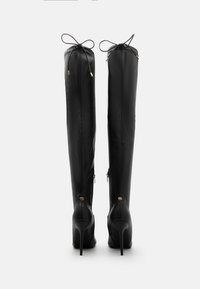 Botas de cuero negras por encima de la rodilla con finos tacones de aguja, detalles de cintas atadas en la parte superior y herrajes en tono dorado en la parte trasera.