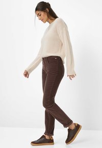 Jeans in corduroy marrone con una vestibilità slim e tasche frontali, abbinati a un maglione lavorato a maglia color crema e sneakers marrone scuro. È visibile una texture morbida.