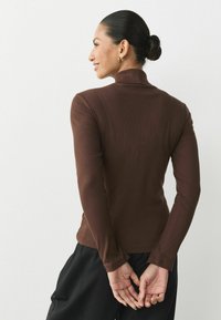 Brown long-sleeve top with a high collar, ribbed texture, and fitted design. Worn with black pants, showcasing a relaxed posture.