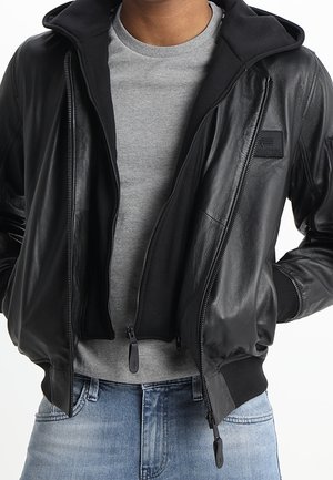 Mand iført en sort læderjakke med hættetrøje over en grå t-shirt og lyseblå denimjeans, hænderne i jakkelommerne.