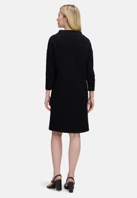 Schwarzes, strukturiertes Kleid mit knielangem Saum, Dreiviertelärmeln und leichtem A-Linien-Schnitt. Getragen mit schwarzen, hohen Sandalen.