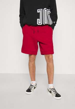 Man draagt rode shorts met trekkoord, zwarte graphic sweatshirt, witte sokken en zwart-witte Nike sneakers, staand met handen in de zakken.
