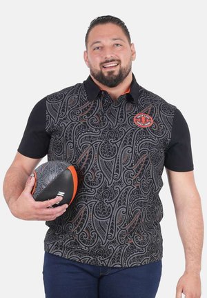 Homme souriant à barbe tenant un ballon de rugby noir et orange, portant un polo noir à motifs avec un logo rond sur la poitrine.