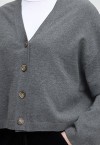 Sivý pletený kardigán s výstrihom do V, päť veľkých okrúhlych gombíkov a rebrovaný lem, vyrobený z mäkkej tkaniny.