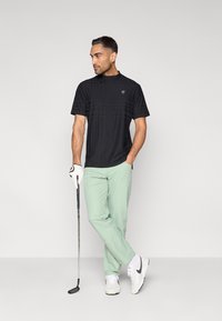 Μαύρο γκολφ μπλουζάκι με κοντά μανίκια και υφή, σε συνδυασμό με ανοιχτό πράσινο παντελόνι. Λευκά αθλητικά παπούτσια και μια ράβδο γκολφ στο χέρι.