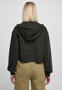 Čierne skrátené mikina s klokankou a kapucňou, s voľnými rukávmi a rebrovanými manžetami. Model má na sebe olivovo zelené nohavice.