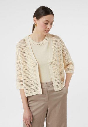 Brede gebreide cardigan in crème met een losse, open structuur, voorzien van halve mouwen en een enkele knoopsluiting. Gedragen over een bijpassend top.