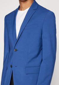Blå blazer med skräddarsydd passform, enkel knappstängning, två framfickor, slät textur och slagkrage. Bärs över en vit skjorta.