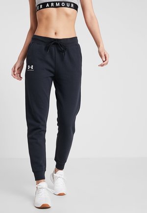 Kobieta ubrana w czarne spodnie dresowe Under Armour i białe trampki, odsłonięty brzuch z biustonoszem sportowym Under Armour na szarym tle.