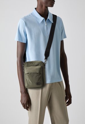 Borsa a tracolla verde in nylon con tasca frontale, accessori in argento e tracolla nera regolabile, indossata sopra una polo azzurra chiara.