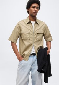 Νέος άνδρας που φοράει μπεζ κοντομάνικο πουκάμισο με φερμουάρ πάνω από λευκή μπλούζα, ανοιχτόχρωμα μπλε τζιν, κρατώντας μαύρο πουλόβερ στο αριστερό χέρι.