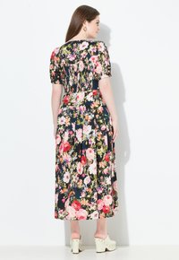 Frau von hinten in einem schwarzen blumenverzierten Midikleid mit kurzen Puffärmeln und weißen, mit Nieten besetzten Plateau-Sandalen.