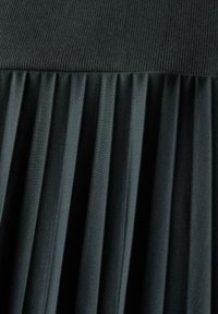 Vêtement noir plissé avec une section supérieure texturée et un tissu lisse en dessous. Présente des plis verticaux pour plus de dimension et de flexibilité.