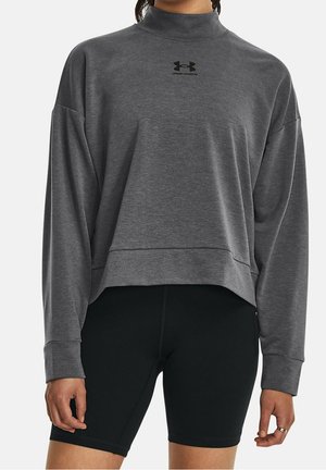 Femme portant un t-shirt gris à manches longues Under Armour et un short noir ajusté, debout devant un fond uni.