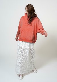 Koraalkleurige oversized hoodie met een kangoeroezak, gecombineerd met een witte maxi rok van kant met bloemenpatronen, en sneakers met paarse accenten.
