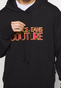 Svart hoodie med "VERSACE JEANS COUTURE" i fet röd och gul text. Inkluderar en dragkedja på huvan och en framficka. Mjuk tygstruktur.