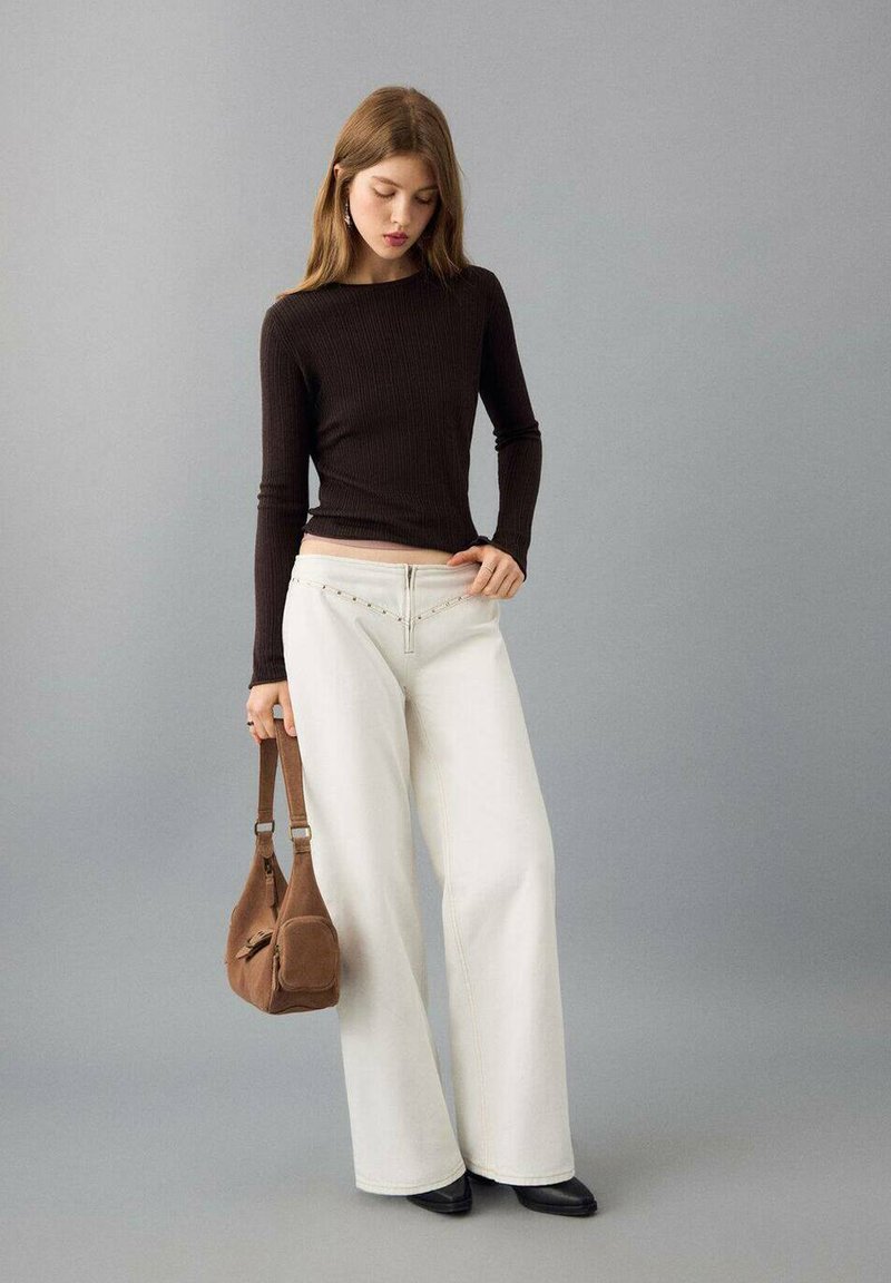 Haut marron à manches longues côtelé, jean blanc à jambes larges avec des coutures décoratives, et un sac à bandoulière en daim marron. Des chaussures noires complètent le look.