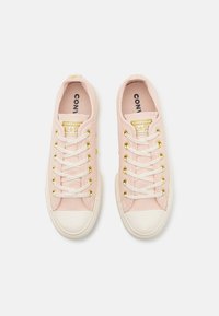 Világos rózsaszín vászon sportcipők, fehér gumi orrbetéttel, arany fűzőlyukakkal és fehér fűzőkkel. Az orrán található a "CONVERSE ALL STAR" logó.